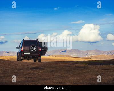 Ein schwarzer SUV steht im goldenen Herbstfeld im Hochland unter einem blauen Himmel mit gelegentlichen weißen Cumuluswolken. Stockfoto