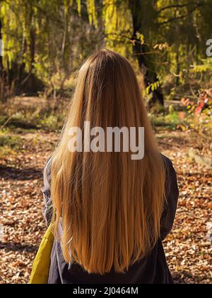 Eine blonde Frau mit langen, fließenden Haaren steht an einem sonnigen Tag im Herbstpark. Rückansicht. Das Konzept der Natürlichkeit Stockfoto