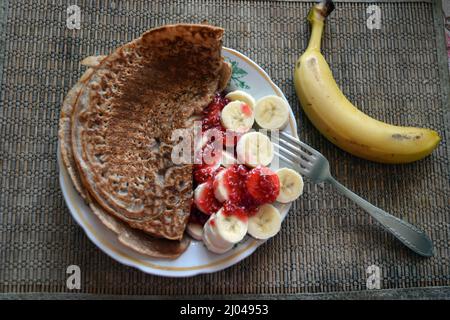 Frisch geschnittene Bananen mit süßer Himbeermarmelade und Schokoladenpfannkuchen auf einer hellen Tonplatte, eine gelbe ganze Banane, ein Küchenmesser und eine Gabel Stockfoto