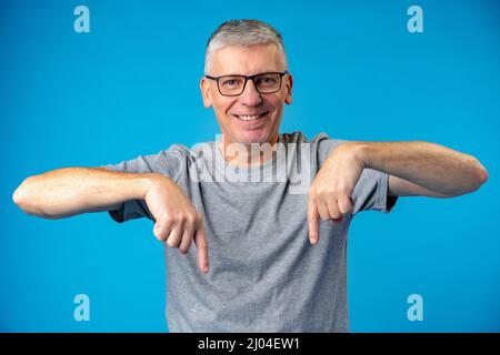 Gutaussehender Mann mittleren Alters, der auf blauem Hintergrund nach unten zeigt Stockfoto