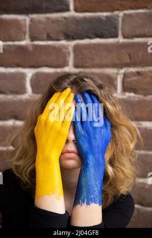 Junge traurige Frau in Verzweiflung bedeckt Gesicht mit Händen in Farben der ukrainischen Flagge gemalt. Krieg in der Ukraine. Leiden der Zivilisten, Schmerzen der Ukrainer. As Stockfoto