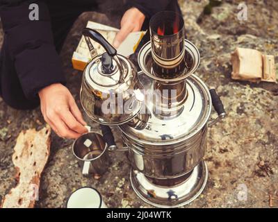 Ein Wasserkocher und ein Samowar mit kochendem Wasser stehen auf Stein, und ein Mann legt Klumpen Zucker in einen Becher. Teezeremonie in der Natur Stockfoto