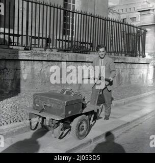 1960s, historisch, Ende des Semesters und ein männlicher Universitätsstudent, in einer Jacke und Krawatte, der auf einem schmalen Bürgersteig neben der Stadtstraße entlang ging, einen metallgerahmten zweirädrigen industriellen Platorm-Trolley schiebt, der seinen Kofferraum und seinen Lederholdall trägt, Oxford, England, Großbritannien. Stockfoto