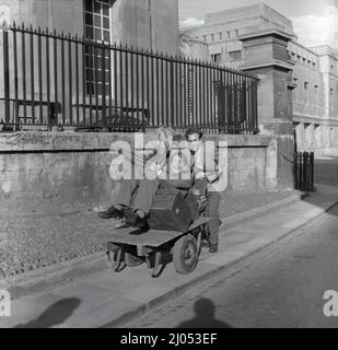 1960s, historisch, Ende des Semesters und ein männlicher Universitätsstudent, in einer Jacke und Krawatte, der auf einem schmalen Bürgersteig neben der Stadtstraße entlang ging, einen metallgerahmten zweirädrigen industriellen Platorm-Trolley schiebt, seinen Kofferraum, einen Holdall und viel zu ihrer Unterhaltung trägt, einer seiner studentischen Freunde, Oxford, England, Großbritannien. Stockfoto