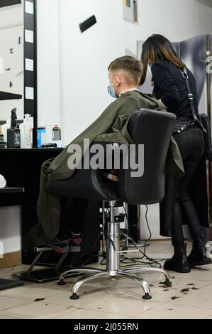 Ein Teenager in einem Schönheitssalon bekommt einen Haarschnitt, ein Friseur schneidet den Jungen eines Teenagers, einen Haarschnitt mit einem Haarschneider und einem Kamm. Stockfoto