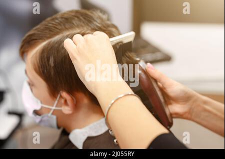 Ein Teenager in einem Schönheitssalon bekommt einen Haarschnitt, ein Friseur schneidet den Jungen eines Teenagers, einen Haarschnitt mit einem Haarschneider und einem Kamm. Stockfoto