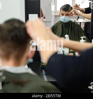 Ein Teenager in einem Schönheitssalon bekommt einen Haarschnitt, ein Friseur schneidet einem Teenager die Haare, einen Haarschnitt mit einer Schere und einem Kamm. Stockfoto