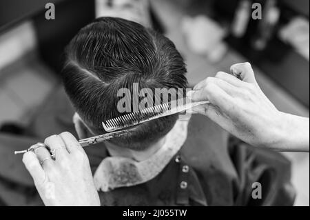 Teen Kerl bekommt einen Haarschnitt während einer Pandemie in einem Friseurladen, Haarschnitt und Trocknen der Haare nach einem Haarschnitt, Haarschnitt mit einer Schere. Stockfoto