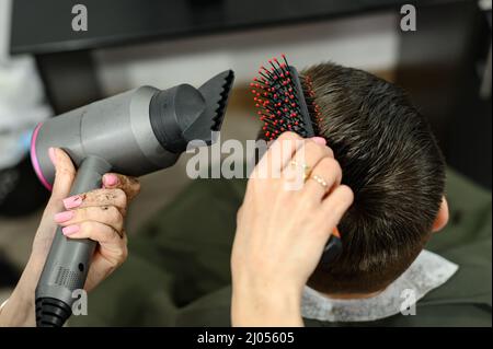 Teenager bekommt einen Haarschnitt während einer Pandemie in einem Friseurladen, einen Haarschnitt und trocknet Haare nach einem Haarschnitt, styling Haare nach einem Haarschnitt mit einem Haartrockner. Stockfoto