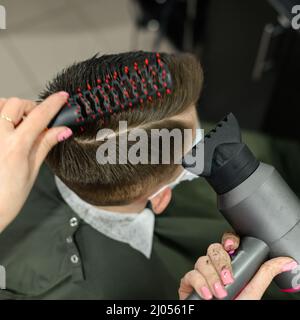 Teenager bekommt einen Haarschnitt während einer Pandemie in einem Friseurladen, einen Haarschnitt und trocknet Haare nach einem Haarschnitt, styling Haare nach einem Haarschnitt mit einem Haartrockner. Stockfoto