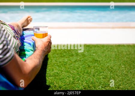 Mittlerer Teil eines Mannes, der sich mit einem Bier in der Hand auf einer Sonnenliege am Pool entspannt. Speicherplatz kopieren Stockfoto