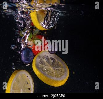 Rote Erdbeeren, gelbe Zitronen und Heidelbeeren fallen ins Wasser. Wasserspritzer, Tropfen. Langsame Bewegung, schwarzer Hintergrund. Stockfoto