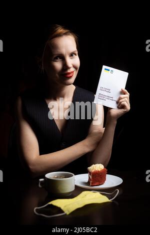 Kiew, Ukraine - 10. November 2021: Eine Frau sitzt bei Tee und Kuchen an einem Tisch und besitzt eine internationale ukrainische Impfbescheinigung. Stockfoto