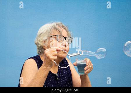 Goiânia, Goias, Brasilien – 16. März 2022: Eine ältere Frau, die Seifenblasen mit blauem Hintergrund macht. Stockfoto