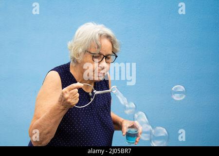 Goiânia, Goias, Brasilien – 16. März 2022: Eine ältere Frau, die Seifenblasen mit blauem Hintergrund macht. Stockfoto