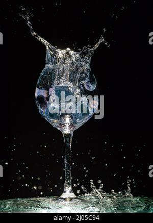 Weinglas vor dunklem Hintergrund mit Eiswürfeln und Heidelbeeren mit vielen Wasserspritzern für Werbung und Gesundheit Stockfoto