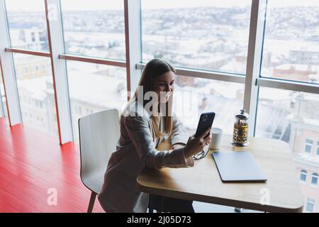 Fokussierte freiberufliche Mitarbeiterin, die in moderner Arbeitsfläche mit offenem Netbook sitzt und Benachrichtigungen auf dem Mobiltelefon in legerer Kleidung überprüft. Stockfoto