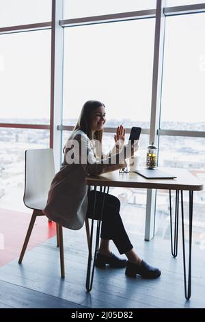 Fokussierte freiberufliche Mitarbeiterin, die in moderner Arbeitsfläche mit offenem Netbook sitzt und Benachrichtigungen auf dem Mobiltelefon in legerer Kleidung überprüft. Stockfoto