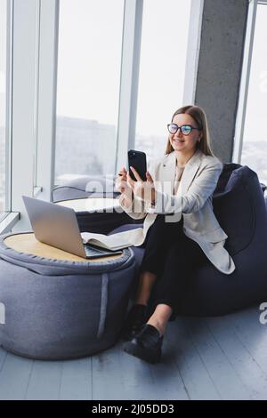 Fokussierte freiberufliche Mitarbeiterin, die in moderner Arbeitsfläche mit offenem Netbook sitzt und Benachrichtigungen auf dem Mobiltelefon in legerer Kleidung überprüft. Stockfoto