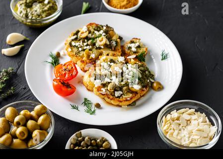 Blumenkohlesteak mit Gewürzen, Chimichurri-Sauce, Mandelflocken, Oliven, gebratenen Kirschtomaten und Kapern auf einem weißen Teller. Dunkler Hintergrund. Stockfoto