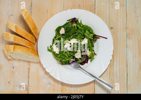 Rucola gemischter Salat mit Gorgonzola-Stücken und Baguette-Scheiben auf Holztisch. Stockfoto