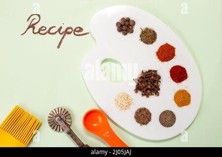 Mehrere Gewürze (Paprika, Kurkuma, alle Gewürze, Nelken, Pfeffer) auf einer weißen Malerpalette, Rezept Wort geschrieben mit brauner Kreide auf hellgrünem Papier Stockfoto
