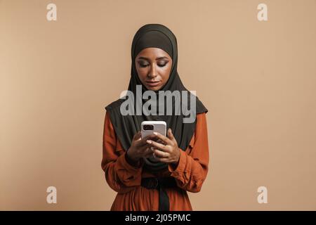 Junge schwarze muslimische Frau in Hijab mit Mobiltelefon isoliert auf beigem Hintergrund Stockfoto