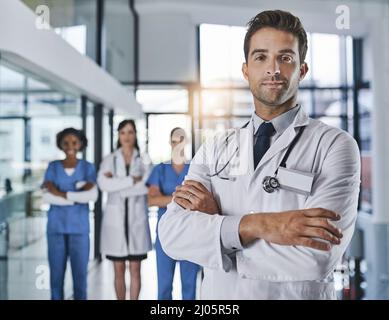 In einem hochleistungsfähigen Krankenhaus. Porträt eines selbstbewussten jungen Arztes, der in einem Krankenhaus arbeitet, mit seinen Kollegen im Hintergrund. Stockfoto
