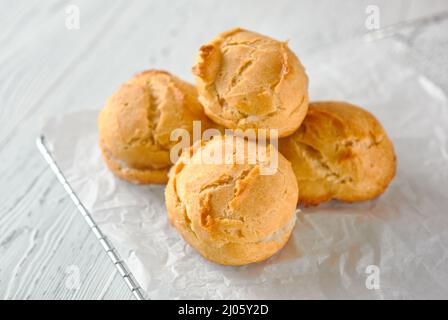 Selbstgemachte Profiterolen auf einem Metallgitter und weißes Papier auf weißem Holzhintergrund. Stockfoto