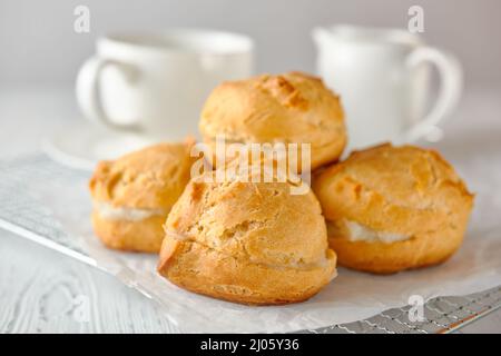 Selbstgemachte Profiterolen auf einem Metallgitter und weißes Papier auf weißem Holzhintergrund. Dahinter befinden sich eine Teetaße und ein Milchkännchen. Stockfoto