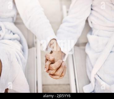 Wir stecken zusammen, meine Liebe. Aufnahme von zwei unkenntlichen Personen, die sich tagsüber in Bademänteln in einem Spa entspannen und die Hände halten. Stockfoto