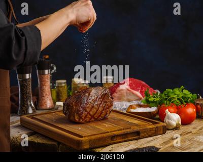 Die Hände des Küchenchefs salzen einen gebackenen Schinken auf einem hölzernen Schneidebrett. Festliches Gericht, Restaurant- und Hausmannsrezepte, Bio-Lebensmittel. Dunkler Hintergrund. Stockfoto