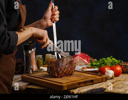 Der Koch schneidet einen saftigen Schinken auf ein Schneidebrett. Gemüse, Kräuter, Gewürze. Dunkler Hintergrund. Restaurant, Hotel, Café, Hausmannskost, Rezepte für Fleisch di Stockfoto