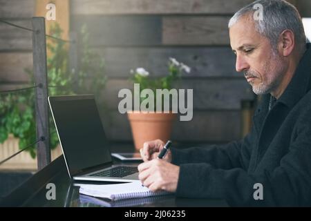 Notizen machen. Aufnahme eines reifen Geschäftsmannes, der Notizen schreibt, während er seinen Laptop in einem Café benutzt. Stockfoto