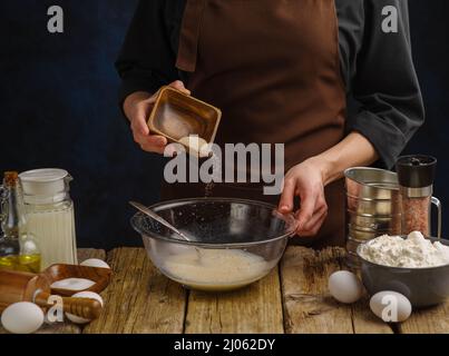 Der Koch bereitet den Teig auf einem Holztisch in einer Glasschüssel zu. Dunkelblauer Hintergrund. Rezepte für Brot, Pizza, Pasta. Restaurant und Hausmannskost. Ad Stockfoto