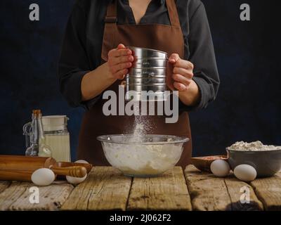 Der Koch bereitet den Teig auf dunklem Grund am Tisch zu. Siebt Mehl durch ein Sieb. Mehl im Schwebezustand. Das Konzept ist die Herstellung von Teig pro Stockfoto