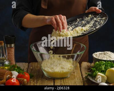 Kochen von Gerichten mit dem Zusatz von geriebenem Käse. Ein professioneller Koch in einer professionellen Küche stellt geriebenen Käse in eine Glasschüssel. Gemüse, Ei Stockfoto