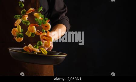 Ein professioneller Koch kocht Garnelen mit Kräutern und Wodka in einer Bratpfanne auf schwarzem Hintergrund. Levitation. Orientalische Küche, Meeresfrüchte, Restaurant, Hotel, Stockfoto