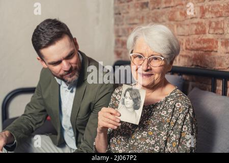 Erinnerungen an die Jugend präsentiert von älteren Inhalten Großmutter im Ruhestand mit einem Foto von ihr im Alter von 20s und sitzt neben ihrem erwachsenen Sohn. Hochwertige Fotos Stockfoto