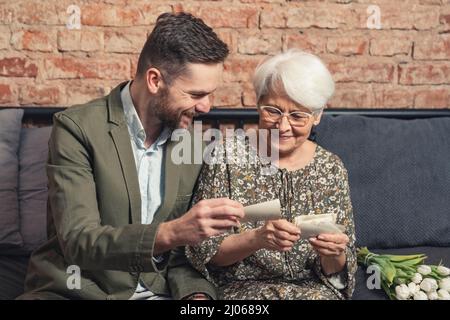 Elegante ältere Rentnerin, die sich mit ihrem erwachsenen Enkelkind verbunden hat, indem sie alte Fotos anschaut und über ihr Leben spricht. Hochwertige Fotos Stockfoto