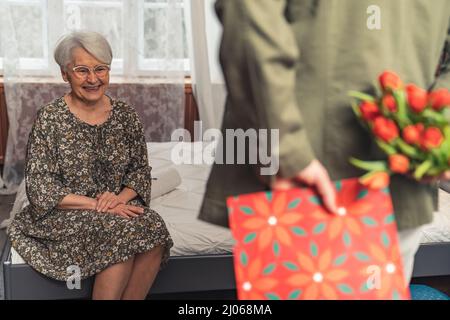 Rückansicht Enkel versteckt Geschenke für seine grinsende grauhaarige kaukasische Rentner Großmutter . Hochwertige Fotos Stockfoto