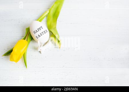 Ein Ei mit Text Frohe Ostern und gelbe Tulpe und weiße kleine Keramik-Hase Stockfoto