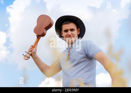 Lustiger Musiker. Junger schöner Kaukasischer, der draußen Ukulele spielt. Schöner Hipster Kerl spielt Ukulele Gitarre auf Natur Hintergrund. Männlich in A Stockfoto