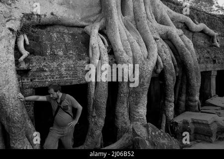 Ein Besucher, der sich am Ta Prohm Tempel, Siem Reap, Kambodscha, an einer riesigen Baumwurzel lehnt (schwarz-weiße Version). Einst als Drehort für einen Hollywood-Film von 2001 verwendet Lara Croft: Tomb Raider mit Angelina Jolie und vor allem bekannt für seine riesigen Wurzeln aus wilden Bäumen, die tief zwischen den Steinen verwurzelt sind, war Ta Prohm ein Kloster für Studenten des Mahayana-Buddhismus, bevor es verlassen und für Hunderte von Jahren vergessen wurde. Stockfoto