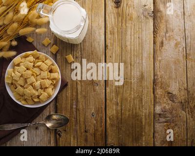 Eine Schüssel Vollkorngetreide, ein Löffel, ein Krug Milch und ein Haufen Getreide auf einem hölzernen Hintergrund. High-Angle-Ansicht. Schnelles Vollkornfrühstück. Weitere Stockfoto