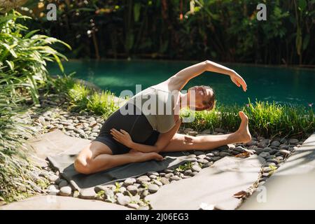 Schwangeren tun Yoga-Posen zu Hause mit Garten auf dem Hintergrund und umarmen ihren Bauch. Sonnenschein, sommerliche Stimmung Stockfoto