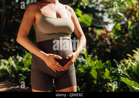 Schwangeren tun Yoga-Posen zu Hause mit Garten auf dem Hintergrund und umarmen ihren Bauch. Sonnenschein, sommerliche Stimmung Stockfoto