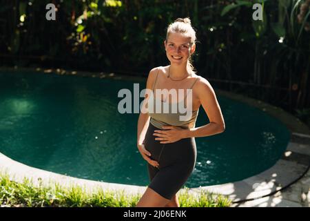 Schwangeren tun Yoga-Posen zu Hause mit Garten auf dem Hintergrund und umarmen ihren Bauch. Sonnenschein, sommerliche Stimmung Stockfoto