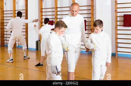 Fokussierte fleißige ernsthafte Jungen fechten aufmerksam dem professionellen freundlichen Fechttrainer im Fitnessstudio zu Stockfoto