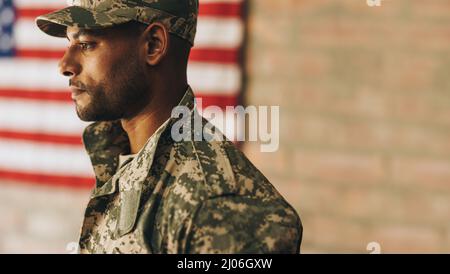 Mutiger junger Soldat, der mit der amerikanischen Flagge im Hintergrund wegschaut. Patriotisches Dienstmitglied in Tarnuniform der Vereinigten Staaten Stockfoto